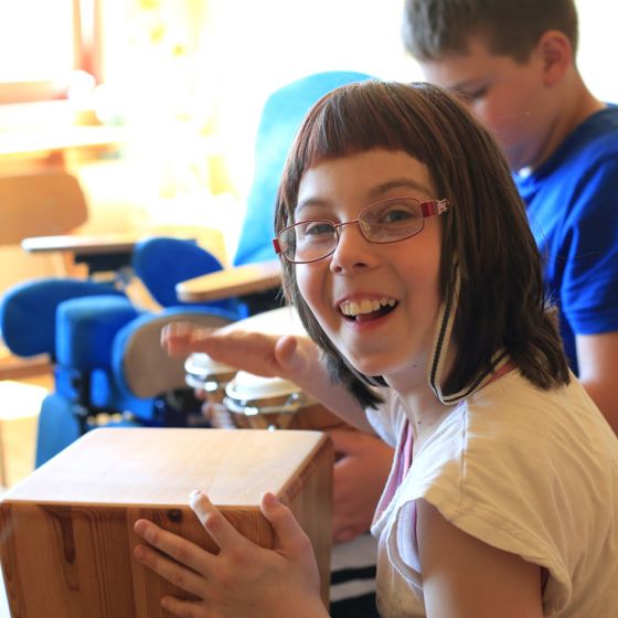 Eine Schülerin mit Brille hält ein Instrument in der Hand im Musikunterricht, schaut lächelnd in die Kamera