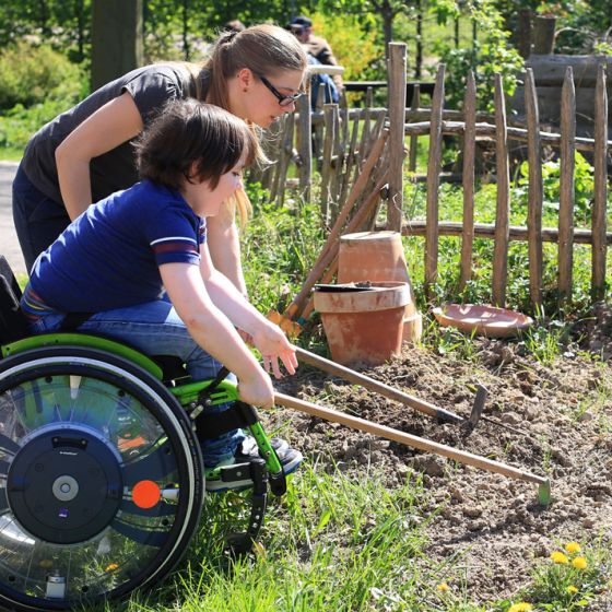 Kind im Rollstuhl arbeitet im Garten mithilfe einer Betreuerin